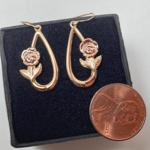 May include: A pair of rose gold-toned teardrop-shaped dangle earrings featuring a sculpted rose flower and leaf design. The earrings are displayed next to a 2023 United States copper-colored penny for scale, showing their relative size.