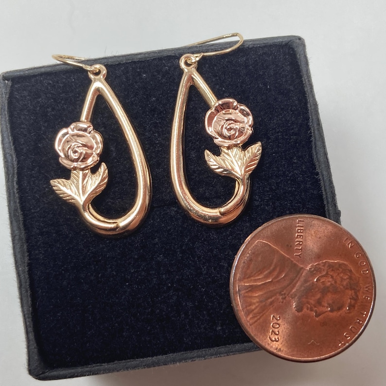 May include: A pair of rose gold-toned teardrop-shaped dangle earrings featuring a sculpted rose flower and leaf design. The earrings are displayed next to a 2023 United States copper-colored penny for scale, showing their relative size.