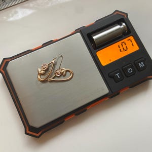 May include: A pair of gold-toned earrings featuring a teardrop-shaped frame with a rose and leaf design at the top. The earrings are placed on a digital scale that displays a weight of 1.07 grams, with a partial ruler visible in the corner.