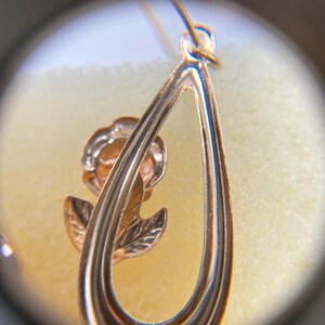 May include: A gold-toned metal earring featuring a teardrop-shaped frame with a rose motif inside. The rose has detailed petals and leaves, and the earring is suspended from a small circular loop at the top, captured through a magnifying lens.
