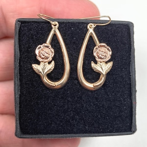 May include: A pair of rose gold-toned drop earrings featuring a teardrop-shaped frame with a sculpted rose flower and leaf design at the base. The earrings are suspended from thin wire hooks and are displayed against a black velvet-textured surface.