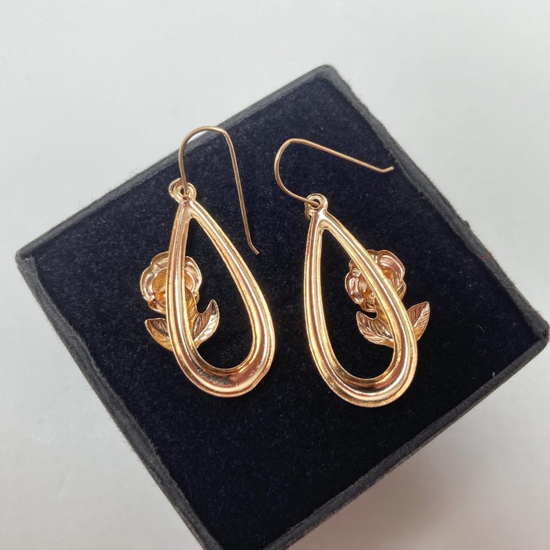 May include: A pair of rose gold-toned drop earrings featuring a teardrop-shaped frame with a small rose flower and leaf motif centered within each loop. The earrings are suspended from curved wire hooks and have a polished metallic finish.