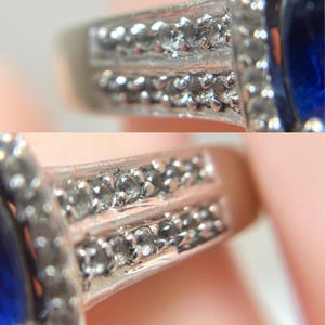 May include: Close-up of a silver ring with a large, oval, deep blue gemstone. The ring's band features two rows of small, clear accent stones. The setting around the blue stone is also adorned with smaller, clear stones, creating a sparkling effect.