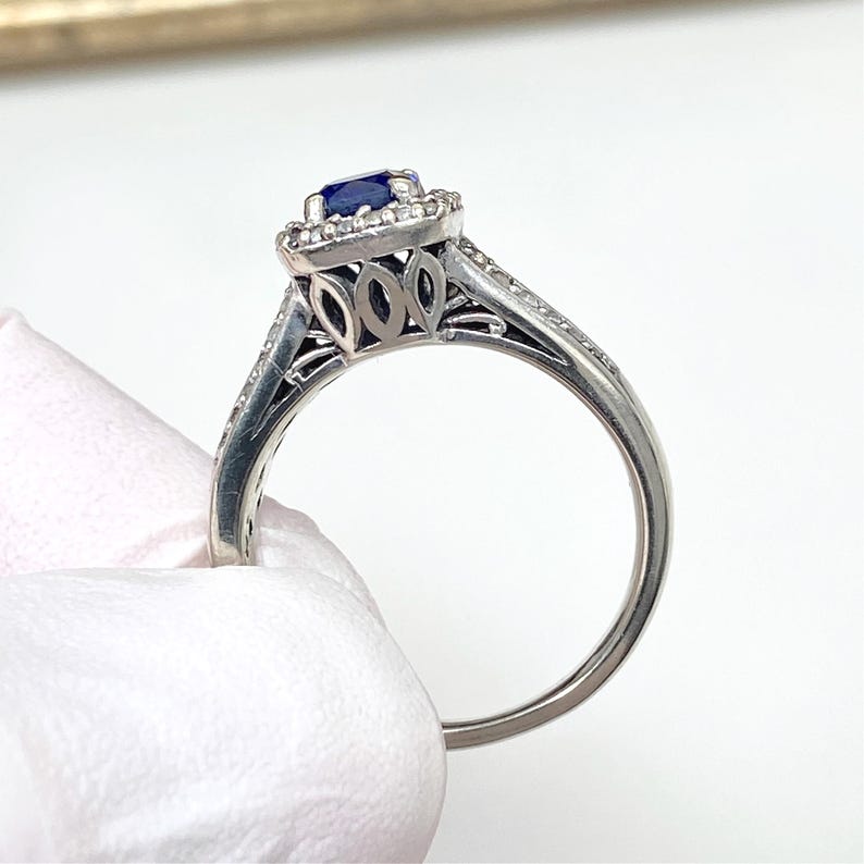 May include: A silver ring with a square, dark blue gemstone center, surrounded by smaller clear stones. The ring has a decorative filigree design on the sides and small clear stones set into the band.