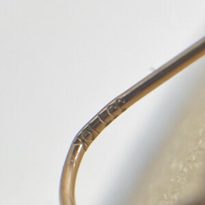 May include: A curved gold earring wire featuring a small, stamped hallmark that reads 10K. The metal has a polished, reflective surface and a smooth, rounded profile, showing the fine detail of the metalwork and the clear engraving of the karat mark.