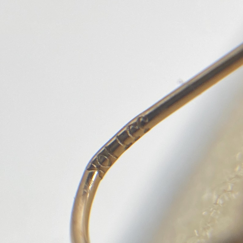 May include: A curved gold earring wire featuring a small, stamped hallmark that reads 10K. The metal has a polished, reflective surface and a smooth, rounded profile, showing the fine detail of the metalwork and the clear engraving of the karat mark.