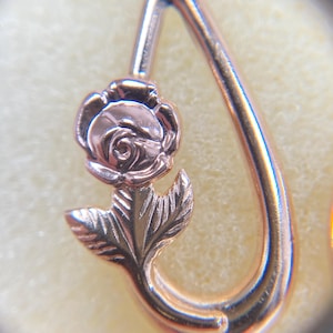 May include: A rose gold-toned metal pendant featuring a sculpted rose flower with detailed petals and leaves. The floral design is integrated into a teardrop-shaped frame, showcasing a polished, reflective surface and intricate metalwork throughout.