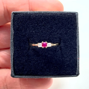 Könnte beinhalten: Ein goldener Ring mit einem zentralen roten Edelstein, flankiert von zwei kleineren weißen Edelsteinen. Der Ring befindet sich in einer dunkelblauen Schmuckschatulle, die das Design und den Farbkontrast hervorhebt.