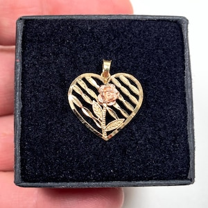 May include: A gold-colored pendant in the shape of a heart with a rose in the center. The heart has a striped design. The pendant is displayed in a black box.