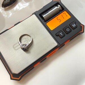 May include: A silver ring with a dark blue gemstone is placed on a digital scale. The scale displays a weight of 5.17. The ring has an ornate design with small, clear stones surrounding the center gemstone. The scale is black and orange.