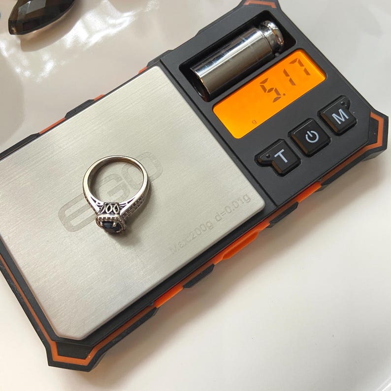 May include: A silver ring with a dark blue gemstone is placed on a digital scale. The scale displays a weight of 5.17. The ring has an ornate design with small, clear stones surrounding the center gemstone. The scale is black and orange.