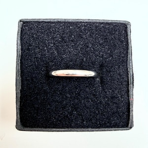 May include: A polished silver-toned band ring with a smooth, rounded surface. The ring is positioned horizontally within a square, black velvet-lined display box, which features a central slot designed to hold the jewelry securely in place.