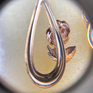 May include: A rose gold-toned metal earring featuring a teardrop-shaped frame that encircles a small, detailed rose flower with a single leaf. The metal has a polished, reflective surface, and the design is captured through a circular lens.