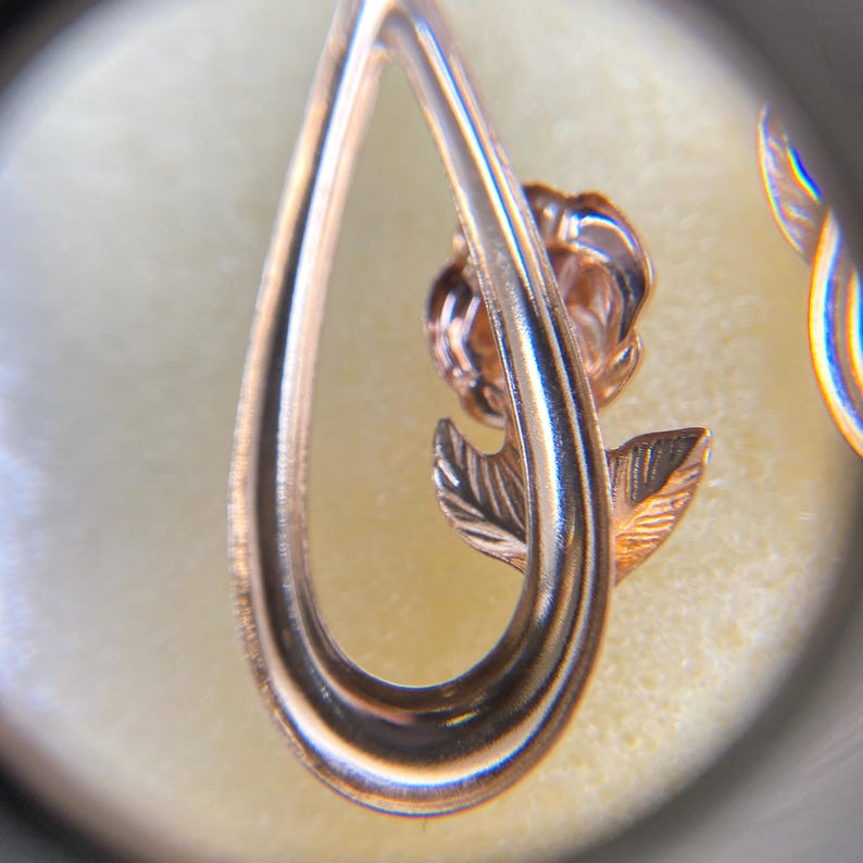 May include: A rose gold-toned metal earring featuring a teardrop-shaped frame that encircles a small, detailed rose flower with a single leaf. The metal has a polished, reflective surface, and the design is captured through a circular lens.