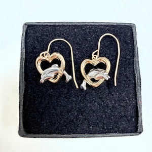 May include: A pair of drop earrings featuring a silver-toned dolphin figure positioned inside a gold-toned heart-shaped frame. Each earring is suspended from a curved gold-toned wire hook, showcasing a two-tone metal design with a polished finish.