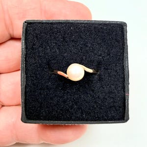 May include: A ring featuring a single round, pale pink pearl set on a polished gold-tone band. The band has a bypass design that curves around the pearl, which is centered within a square black velvet-lined jewelry box for display.