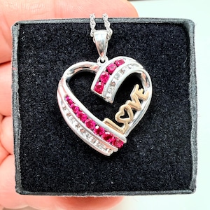 Könnte beinhalten: Eine silberfarbene herzförmige Anhängerkette mit einer Reihe leuchtend rosa runder Steine und dem Wort LOVE in goldfarbenen Buchstaben. Der Anhänger hängt an einer silberfarbenen Kette und liegt in einer mit schwarzem Samt ausgekleideten Box.
