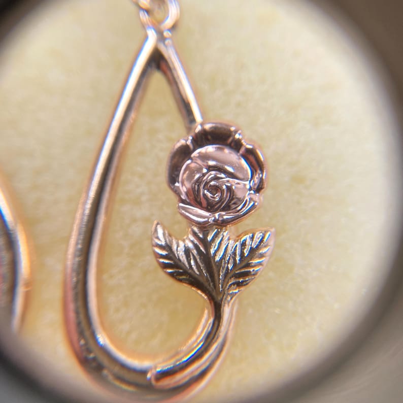 May include: A rose gold pendant featuring a teardrop-shaped frame with a sculpted rose flower and two leaves centered within the open space. The metal has a polished finish, highlighting the detailed petals and foliage of the floral design.