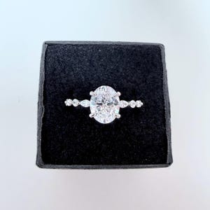 May include: A silver-toned ring featuring a large, central oval-cut clear stone held by four prongs. The band is accented with smaller, alternating marquise-cut and round-cut clear stones extending outward from the center setting.
