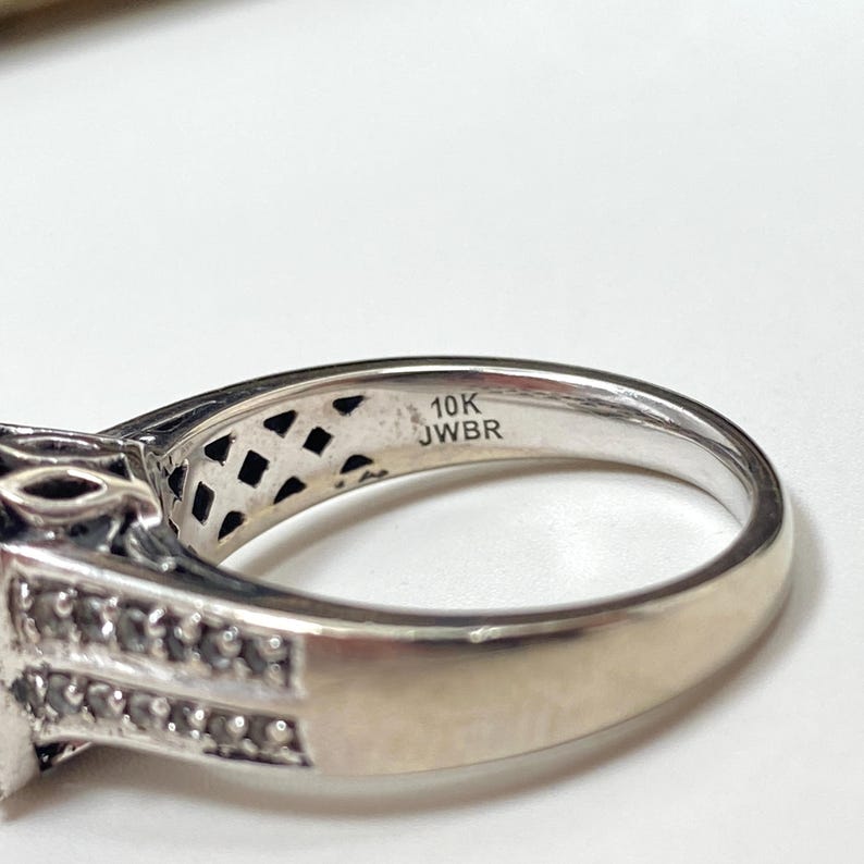 May include: Close-up of a silver ring with a decorative design. The ring features a band with the text "10K JWBR" and a detailed setting with small stones. The ring is set against a white background.