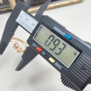 May include: A gold-toned earring featuring a rose motif being measured by a black digital caliper. The digital display clearly shows a measurement of 0.93 inches, indicating the size of the jewelry piece as it is held within the caliper jaws.