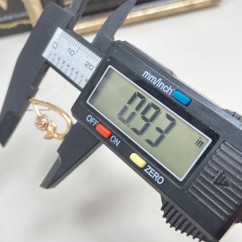 May include: A gold-toned earring featuring a rose motif being measured by a black digital caliper. The digital display clearly shows a measurement of 0.93 inches, indicating the size of the jewelry piece as it is held within the caliper jaws.