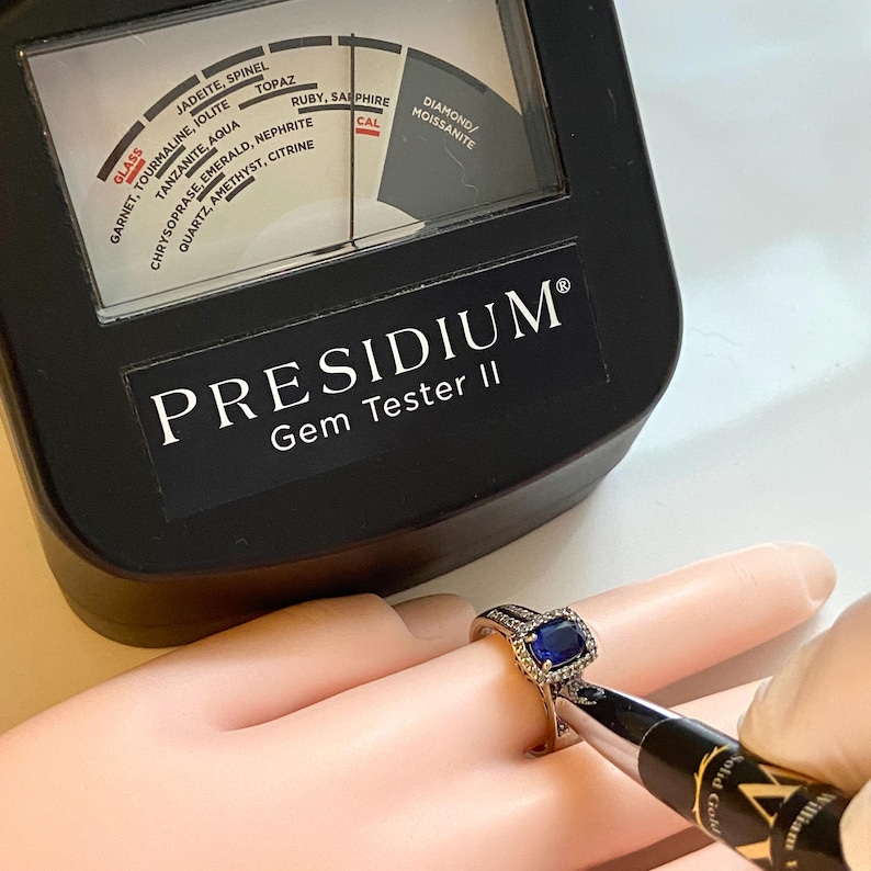 May include: A Presidium Gem Tester II is used to test a ring with a blue gemstone. The tester has a dial with labels for various gemstones, including diamond, ruby, and sapphire. The ring is silver with a square blue stone and small surrounding stones.