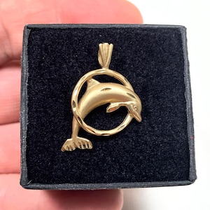 May include: A gold-colored dolphin pendant. The dolphin is depicted leaping through a circular hoop. The pendant is displayed on a black velvet surface, showcasing the jewelry's design and color. The pendant is likely made of gold.