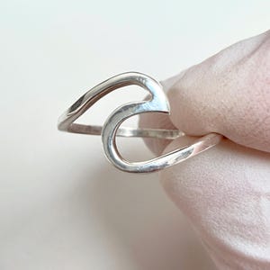 Puede incluir: Un anillo de plata pulida con un diseño fluido y continuo en forma de onda que forma un bucle abierto. El metal tiene una superficie lisa y reflectante con un brillo metálico, mostrando una silueta curva y abstracta alrededor del dedo.