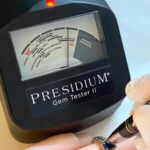 May include: A Presidium Gem Tester II device is used to test a ring with a blue gemstone. The device has a dial with gemstone names like ruby, sapphire, and diamond. The device is black with white text.