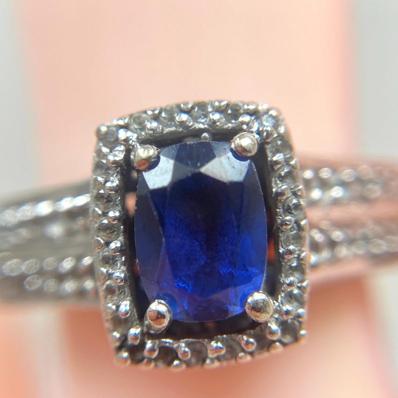 May include: A silver ring featuring a large, oval, deep blue gemstone, surrounded by a halo of smaller, clear stones. The band has a textured design. The ring is set against a soft, blurred background.