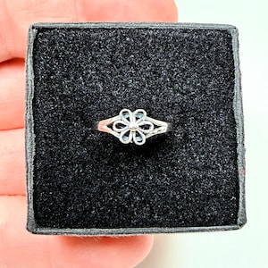 May include: A silver-toned ring featuring a central flower motif with six open petals and a small spherical accent in the middle. The ring is displayed centered on a square black foam insert within a small, dark-colored jewelry box.