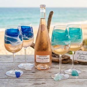 May include: A clear glass wine bottle of Gerard Bertrand Cote des Roses rose wine positioned between four wine glasses. Two glasses feature blue and white resin waves, while two feature teal and white resin waves. Each glass has a small sea glass charm.