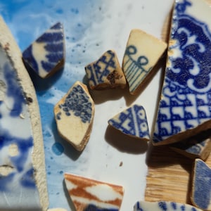 Puede incluir: Primer plano de varios trozos de cerámica rotos con patrones azules y blancos. Los fragmentos muestran varios diseños, incluyendo motivos florales y geométricos, sobre un fondo color crema. Algunas piezas muestran un borde marrón.