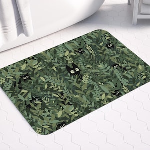 Può includere: Un tappetino da bagno rettangolare con un motivo a fogliame verde e facce di gatti neri nascoste. Il tappetino è posto davanti a una vasca da bagno bianca. Il testo "CHARMEDVILLE" è visibile.