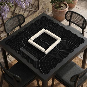 Pu&ograve; includere: Un tappetino da tavolo quadrato nero con un motivo a linee topografiche bianche. Il tappetino &egrave; posizionato su un tavolo nero e presenta una disposizione quadrata di tessere rettangolari bianche al centro, creando un setup di gioco.