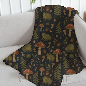 May include: A dark blanket featuring a repeating pattern of green frogs, red-spotted mushrooms, ferns, acorns, keys, and small flowers. The blanket is draped over a grey sofa, displaying the whimsical design.