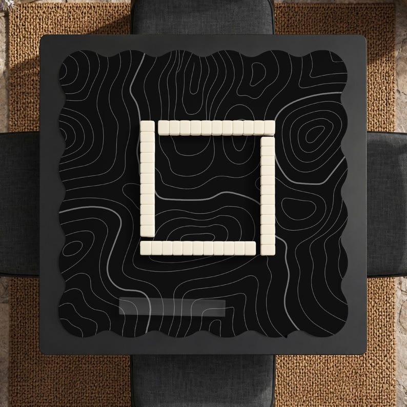 Pu&ograve; includere: Un tappetino da gioco quadrato nero con bordo smerlato e un motivo a linee topografiche bianche. Al centro del tappetino, quattro file di tessere rettangolari color bianco sporco formano una cornice quadrata, con nove tessere per lato.