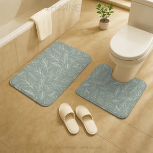 May include: A bathroom set with two sage green bath mats with a white leaf pattern, a pair of cream-coloured slippers, and a white towel. The mats are rectangular and U-shaped, designed to fit around a toilet.