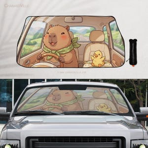 May include: Car windshield sunshade featuring a cartoon capybara driving a car, wearing a green scarf, and a yellow chick passenger. The interior of the car is beige, with a scenic background. The text "www.charmedville.com" is visible.