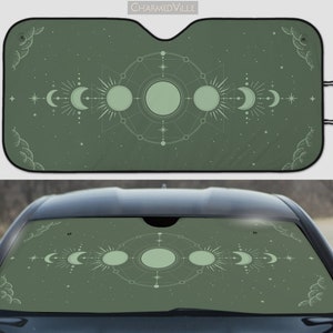May include: A green car sunshade with a white design of a celestial moon and sun phases with geometric shapes and stars. The sunshade is folded and displayed on a car windshield.
