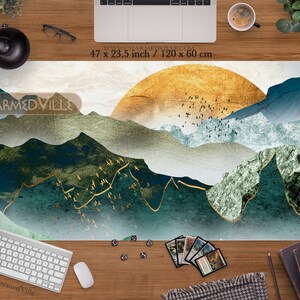May include: A large, rectangular, white and gold gaming mat with a blue and green mountain landscape design. The mat features a large, golden sun in the background and a flock of birds flying over the mountains. The text "CHARMED VILLE" is printed in the top left corner of the mat. The mat measures 47 inches by 23.5 inches.