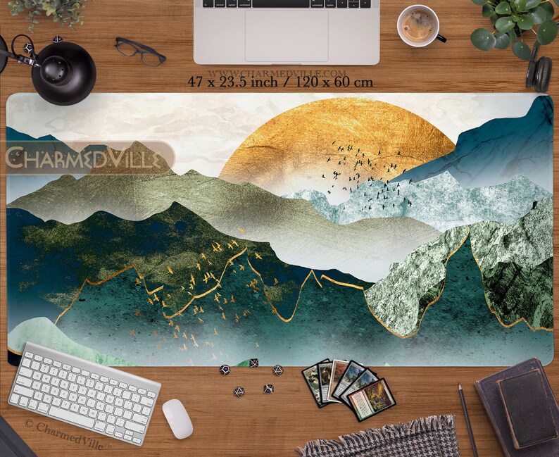 May include: A large, rectangular, white and gold gaming mat with a blue and green mountain landscape design. The mat features a large, golden sun in the background and a flock of birds flying over the mountains. The text "CHARMED VILLE" is printed in the top left corner of the mat. The mat measures 47 inches by 23.5 inches.