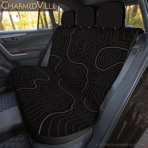 Peut inclure: Housse de si&egrave;ge de voiture noire avec un motif de carte topographique blanc. La housse est install&eacute;e dans le si&egrave;ge arri&egrave;re d'un v&eacute;hicule. La marque "CharmedVille" est visible en haut de l'image.