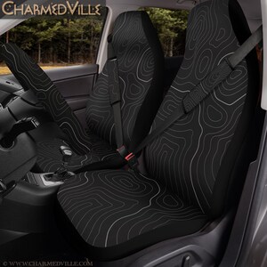 Peut inclure: Housses de si&egrave;ge de voiture noires avec un motif de carte topographique blanc. Les housses sont install&eacute;es dans un v&eacute;hicule, avec l'appui-t&ecirc;te et le dossier. La ceinture de s&eacute;curit&eacute; est visible. Le texte "CHARMEDVILLE" est en haut de l'image.