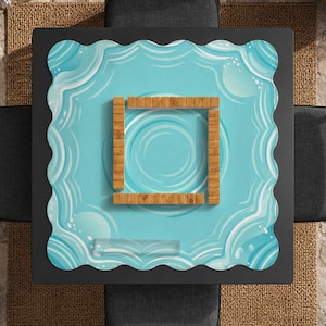 May include: A square, black coffee table with a light blue, wavy-edged surface. The surface features a water-like design with a wooden square in the center. The table is surrounded by gray cushions and a woven rug.
