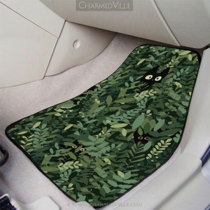 May include: Black cat peeking out from behind green leaves on a car floor mat. The mat has a black border and the text "CHARMEDVILLE" is visible at the top of the image.
