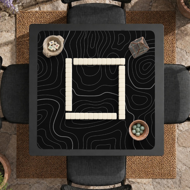 Pu&ograve; includere: Un tavolo da gioco quadrato con un tappetino nero caratterizzato da un motivo a linee topografiche bianche. Al centro &egrave; disposta una serie quadrata di tessere mahjong bianche. Una ciotolina con dadi e una con sfere verdi sono posizionate.