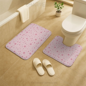 Könnte beinhalten: Ein Badezimmer-Set mit einer rechteckigen Badematte und einer U-förmigen Toilettenmatte, beide in Hellrosa mit einem Blumenmuster. Zwei cremefarbene Hausschuhe liegen auf dem Boden. Ein weißes Handtuch hängt am Rand einer Badewanne.