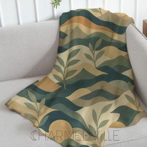 May include: A patterned throw blanket in shades of green, tan, and gold, with a design of rolling hills and leaf sprigs. The blanket is draped over a light grey sofa. The text "CHARMEDVILLE" is visible. The blanket's design evokes a natural landscape.